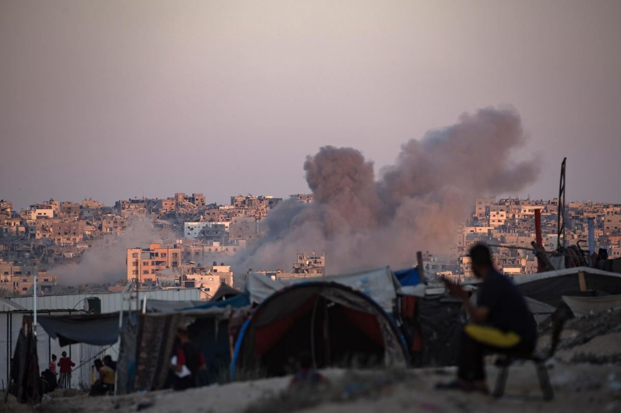 El humo se eleva tras un ataque aéreo israelí mientras los palestinos desplazados internos se sientan junto a sus tiendas de campaña en el campamento de Khan Younis, al sur de la Franja de Gazal. (Foto de Haitram Imad de la agencia EFE)