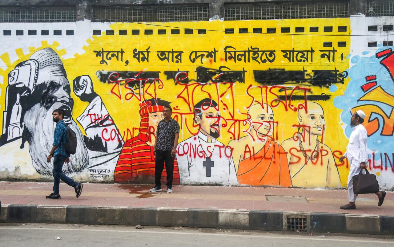 Varias personas caminan junto a un mural en una calle de Daca, este lunes. (Foto de Monirul Alam de la agencia EFE)