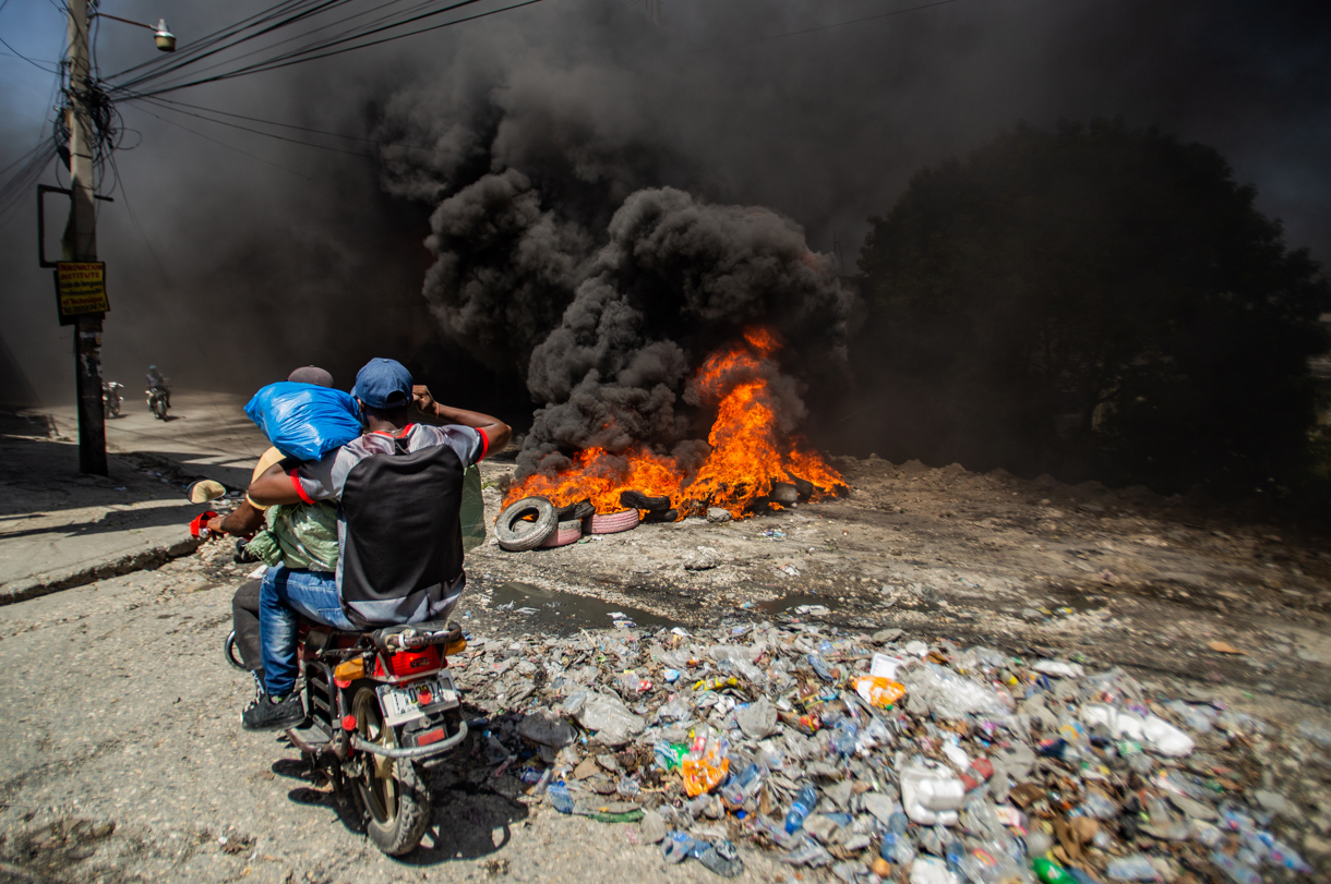 Dos hombres se movilizan en motocicleta frente a una barricada con fuego durante una protesta este lunes, en el barrio Solino, en Puerto Principe (Haití). (Foto de Mentor David Lorens de la agencia EFE)